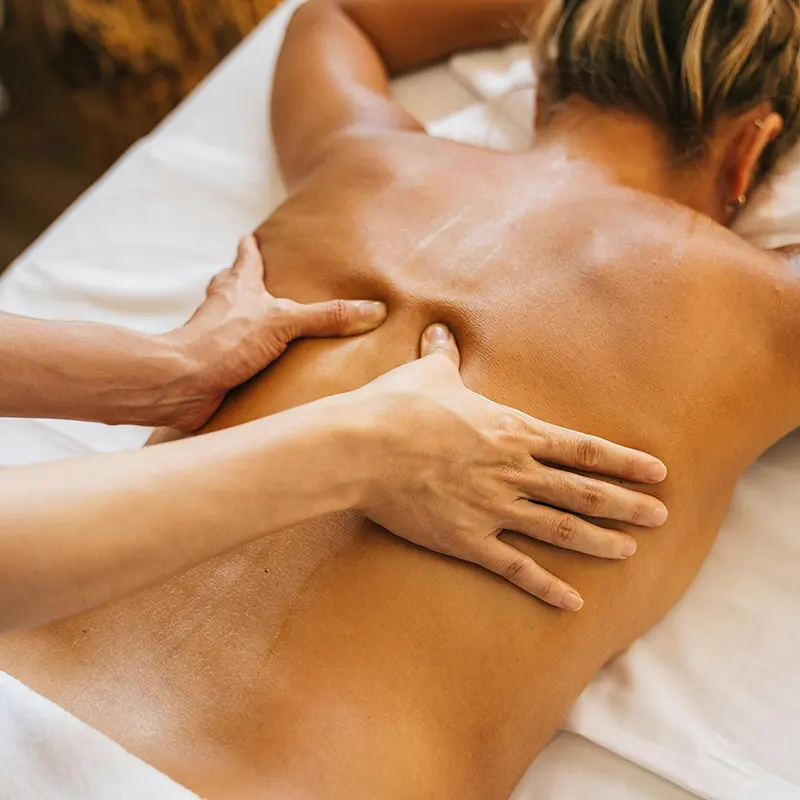 Person receiving a professional back massage while lying face down on a white massage table, with a therapist using both hands to apply pressure along the spine